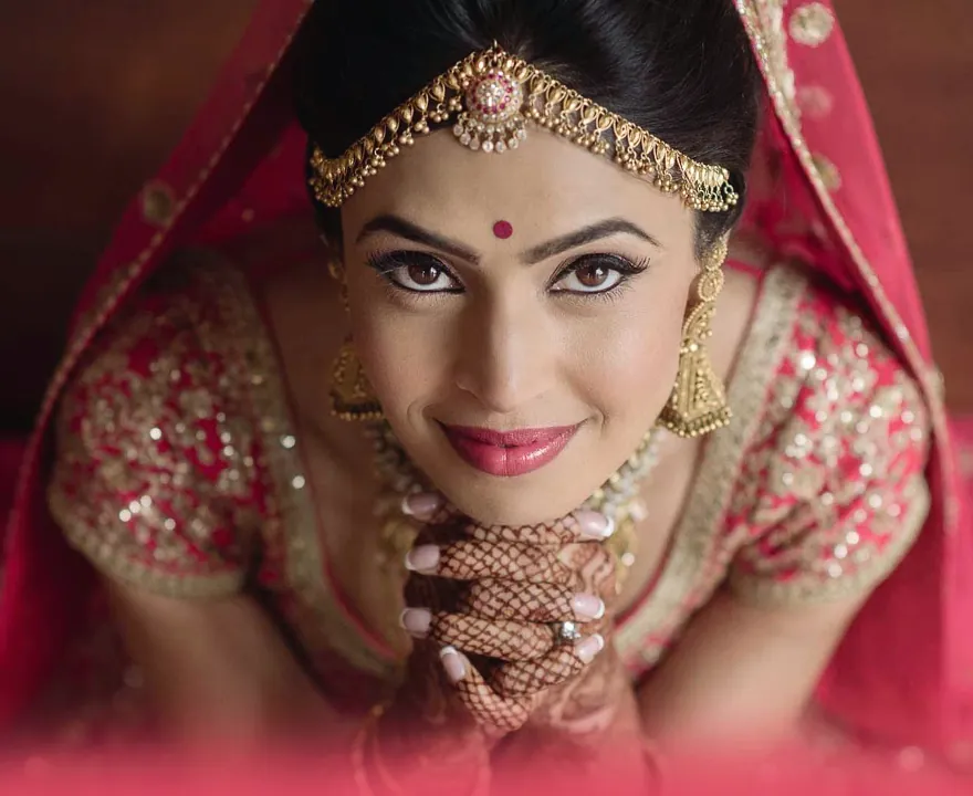 A bride in a red and gold lehenga looking up over mehendi-covered hands, wearing a maang tikka and jhumkas.